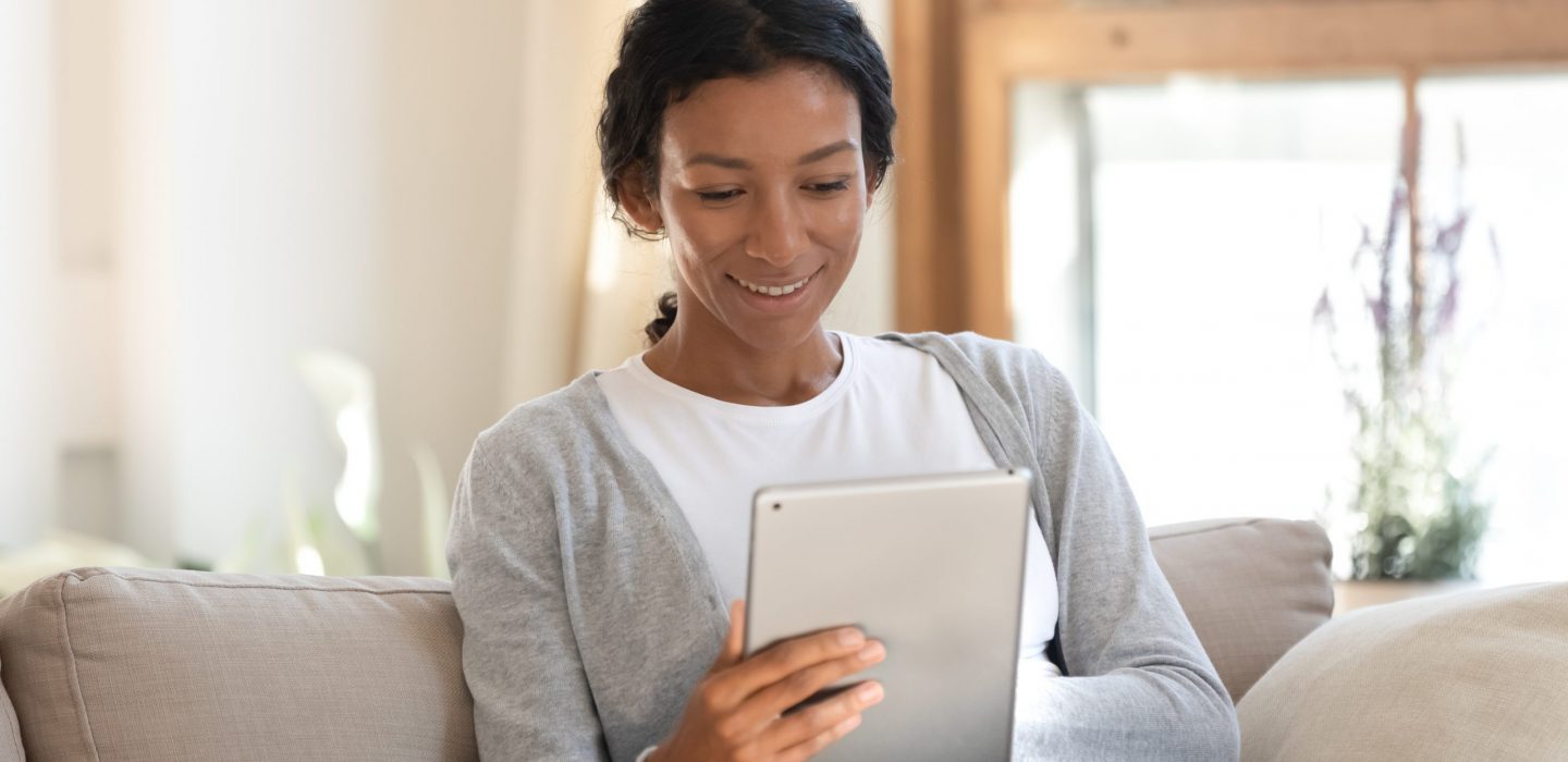 Smiling biracial young female sit relax on sofa at home browsing wireless Internet on tablet, happy African American millennial woman rest on couch in living room watch video or surfing on pad gadget